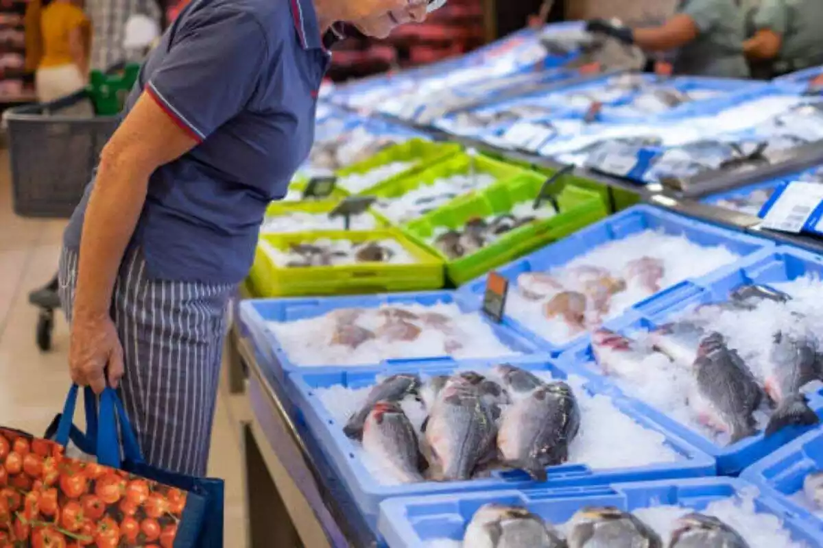 Foto de una pescadería de supermercado con una mujer mirando el pescado que se expone en cajas azules. De fondo, en cajas verdes