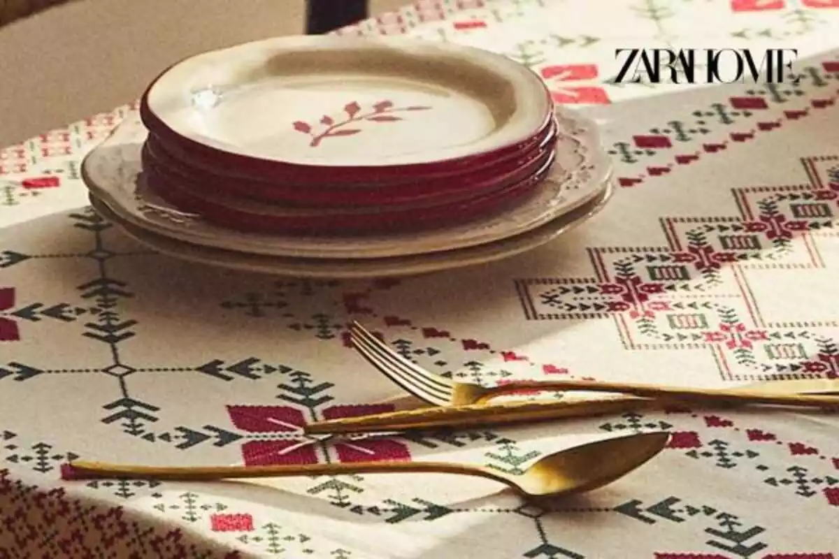 Foto de parte de una mesa con mantel navideño de flores rojas y verdes y unos platos y al lado cubiertos dorados y arriba logo de Zara Home