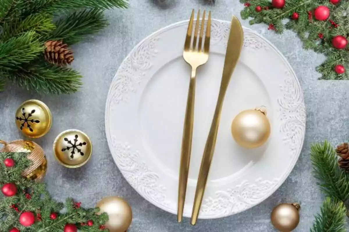Foto de plato de color blanco con cubiertos dorados y cascabeles a los lados y bolas de navidad doradas y en los bordes decoración de acebo y pino